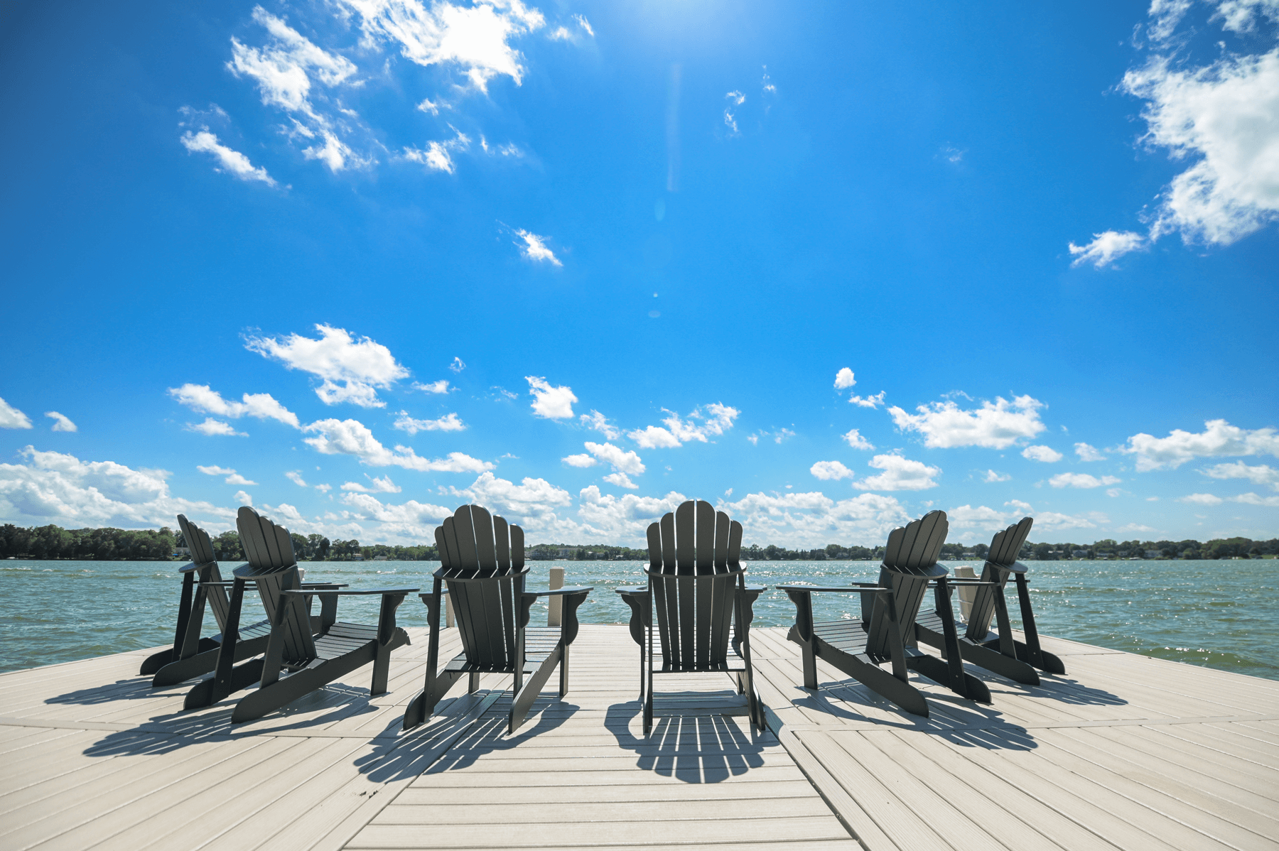 4 adirondack chairs on a dock looking at a lake and blue sky